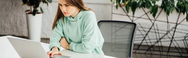 Panoramic shot of upset girl sitting at table and using laptop at home — Stock Photo