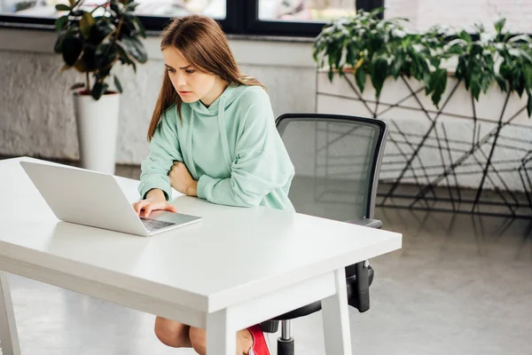 Upset girl in hoodie sitting at table and using laptop at home — Stock Photo