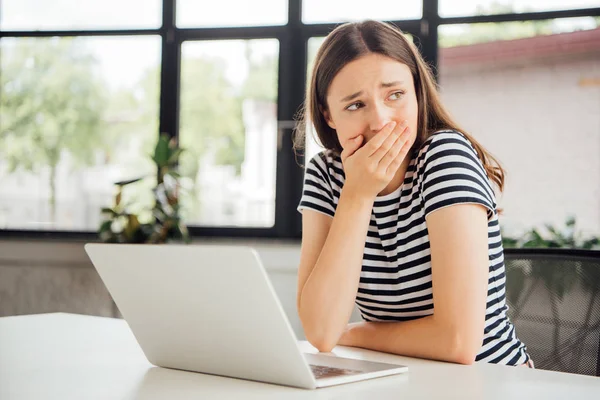 Triste fille en t-shirt rayé couvrant la bouche avec la main tout en utilisant un ordinateur portable à la maison — Photo de stock