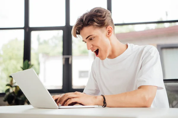 Animado teen menino em branco t-shirt usando laptop em casa — Fotografia de Stock