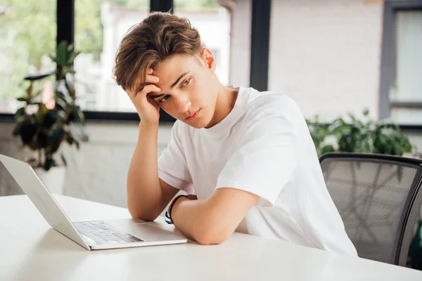 Pensive teen boy in white t-shirt sitting at table with laptop and looking away at home — Stock Photo