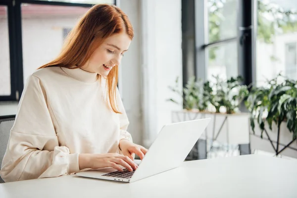 Lächelndes attraktives Mädchen, das am Tisch sitzt und zu Hause Laptop benutzt — Stockfoto