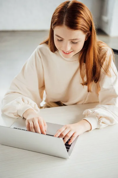 Улыбающаяся привлекательная девушка сидит за столом и использует ноутбук дома — стоковое фото