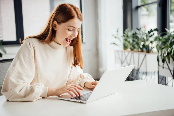 Lächelndes attraktives Mädchen, das am Tisch sitzt und zu Hause Laptop benutzt — Stockfoto