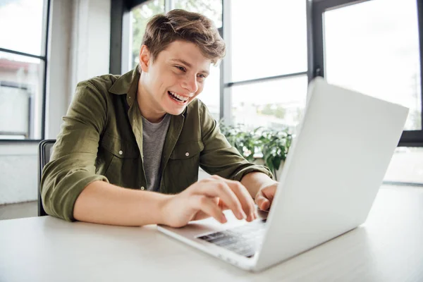 Aufgeregter Teenager sitzt am Tisch und tippt zu Hause auf der Laptop-Tastatur — Stockfoto