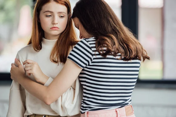 Adolescente chica en rayas camiseta abrazando y apoyando triste amigo - foto de stock