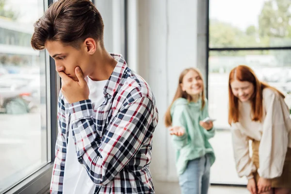 Garçon triste en chemise à carreaux debout près de la fenêtre tandis que les filles se moquent de lui — Photo de stock