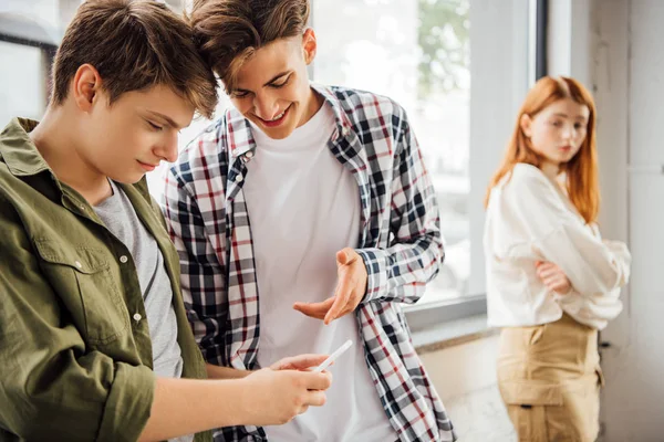 Dos adolescentes felices sonriendo mientras usan el teléfono inteligente en la escuela - foto de stock