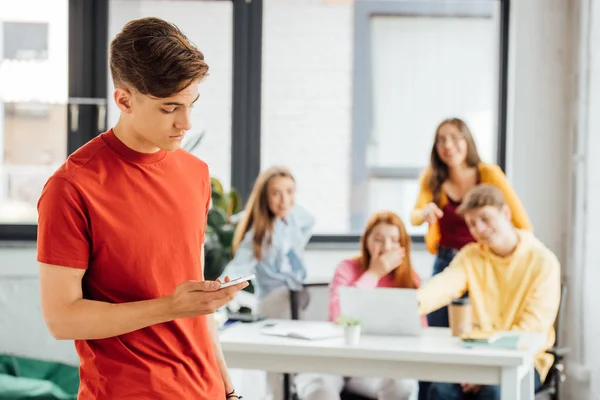 Groupe d'écoliers avec ordinateur portable riant de garçon triste tenant smartphone — Photo de stock