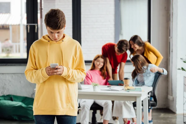 Сумний хлопчик в жовтому светрі використовує смартфон і сміється підлітків за столом — стокове фото