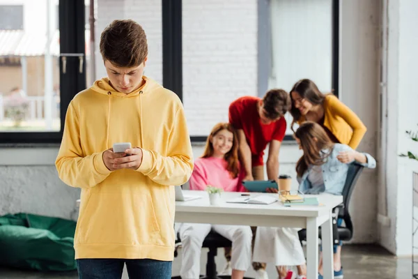 Menino triste com capuz amarelo usando smartphone e rindo adolescentes na mesa — Fotografia de Stock