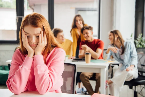 Triste chica sentado en frente de riendo compañeros de clase - foto de stock