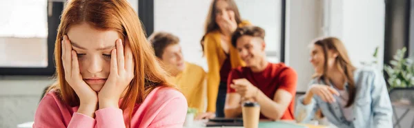 Panoramic shot of schoolchildren laughing while bullying sad girl on foreground — Stock Photo