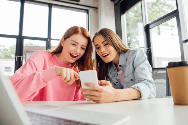 Dos chicas emocionadas sonriendo mientras usan el teléfono inteligente en la escuela - foto de stock