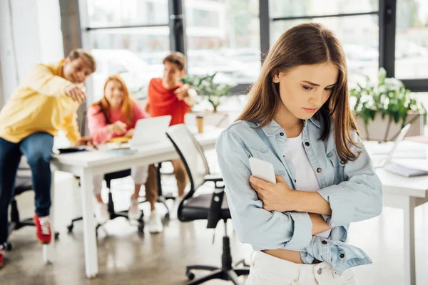 Laughing schoolchildren bullying sad girl with smartphone in school — Stock Photo