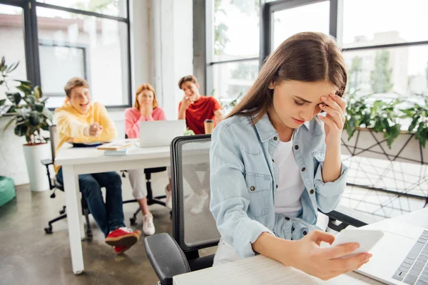 Laughing schoolchildren bullying sad girl with smartphone in school — Stock Photo