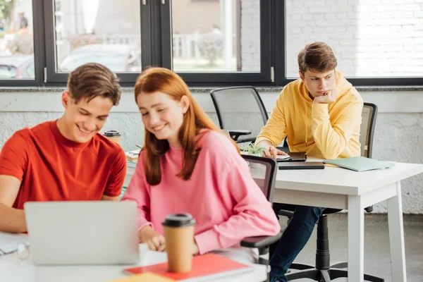 Selective focus of two friends smiling while using laptop and sad boy at desk in school — Stock Photo