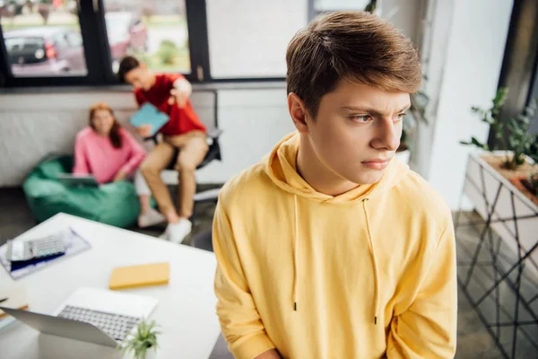 Nachdenklicher Junge im gelben Kapuzenpulli und lachende Mitschüler, die ihn mobben — Stockfoto