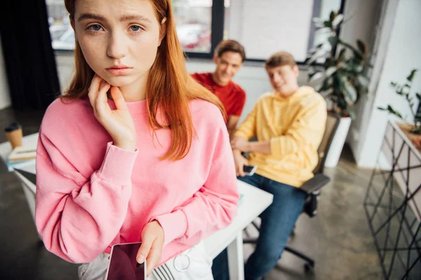 Sad girl holding smartphone while classmates laughing at her in school — Stock Photo