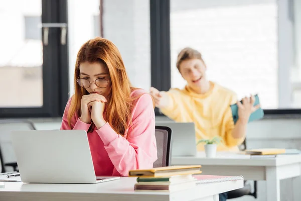 Menina pensativa em óculos usando laptop enquanto colega de classe rindo dela — Fotografia de Stock