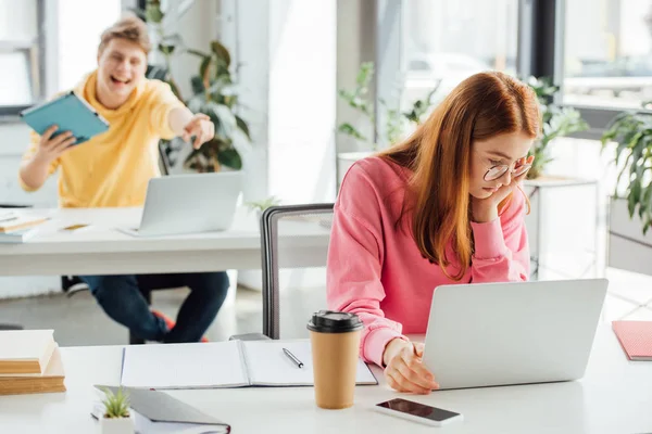 Pensive girl in glasses using laptop while classmate laughing at her — Stock Photo