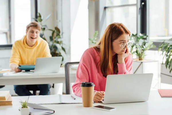 Chica pensativa en gafas usando portátil mientras compañero de clase se ríe de ella - foto de stock