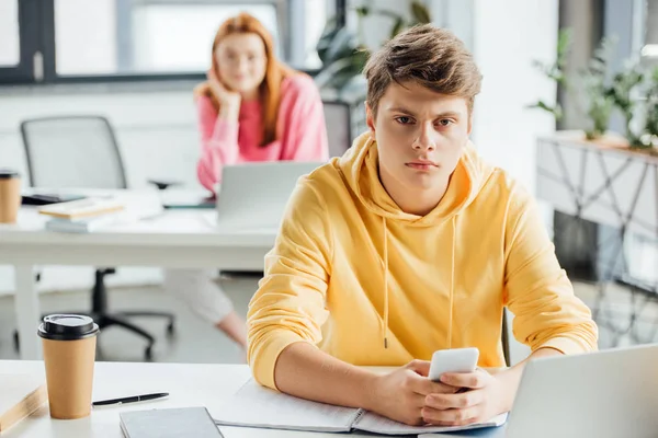 Pensive boy in yellow hoodie using smartphone at desk — Stock Photo