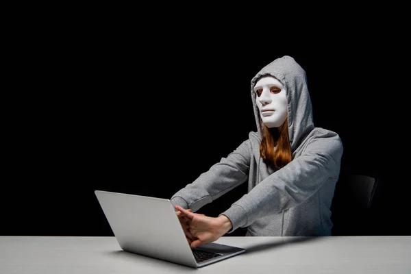 Anonymous girl in mask and hoodie sitting near laptop and stretching hands during cyberbullying isolated on black — Stock Photo