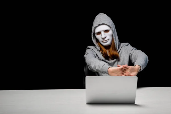 Anonymous girl in mask and hoodie sitting near laptop and stretching hands during cyberbullying isolated on black — Stock Photo