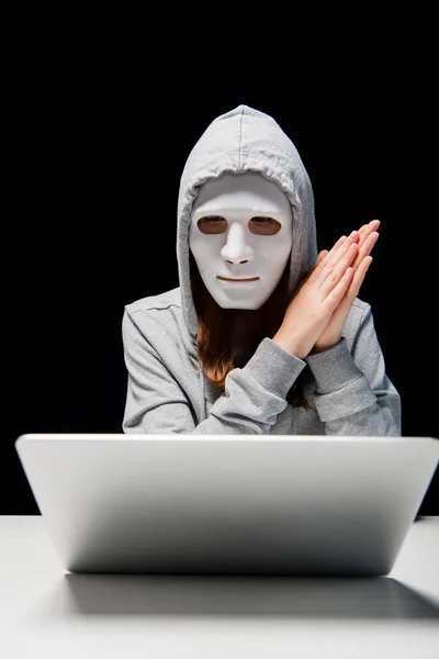 Vista frontal da menina anônima em máscara e capuz sentado perto de laptop e esfregando as mãos durante cyberbullying isolado em preto — Fotografia de Stock