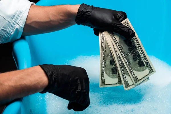 Vista ritagliata dell'uomo in guanti di gomma nera in possesso di banconote dollaro vicino bolle di sapone con acqua — Foto stock