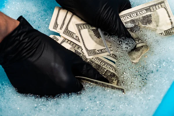 Обрезанный вид человека в черных резиновых перчатках, стирающих долларовые купюры в мыльных пузырях с водой — стоковое фото