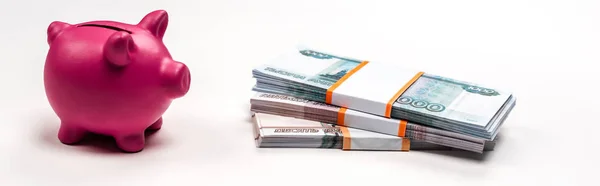 Panoramic shot of pink piggy bank near stacks with russian money on white — Stock Photo