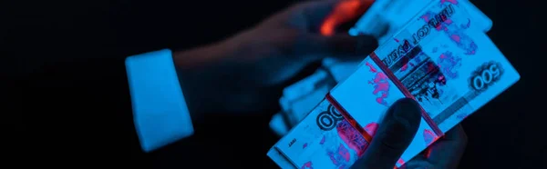 Panoramic shot of man holding russian money under blue uv lighting isolated on black — Stock Photo