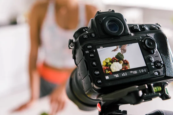 Selective focus of digital camera with sportswoman gesturing near vegetables on screen — Stock Photo