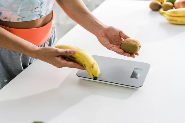Cropped view of woman holding banana and kiwi near food scales — Stock Photo