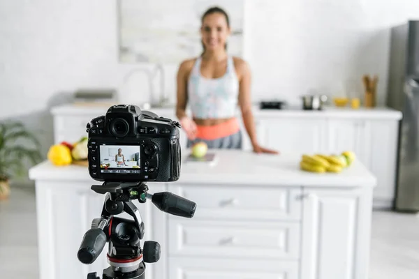 Selective focus of digital camera with happy woman gesturing near vegetables and fruits on screen — Stock Photo