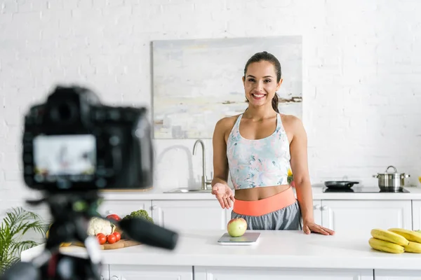 Foco seletivo de menina feliz gesticulando perto de frutas e olhando para a câmera digital — Fotografia de Stock