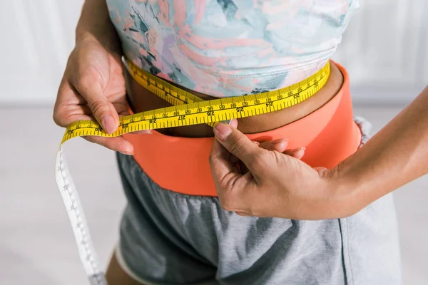 Cropped view of sportive woman holding yellow measuring tape while measuring waist — Stock Photo