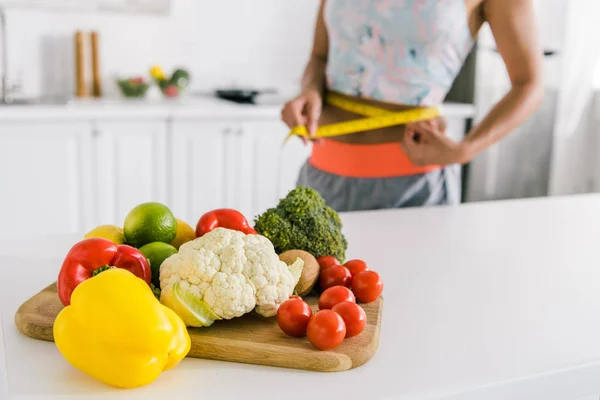 Selective focus of vegetables on cutting board near woman measuring waist — Stock Photo