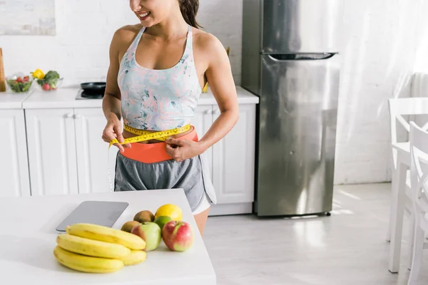 Foco seletivo de desportista feliz medir a cintura perto de frutas na mesa — Fotografia de Stock