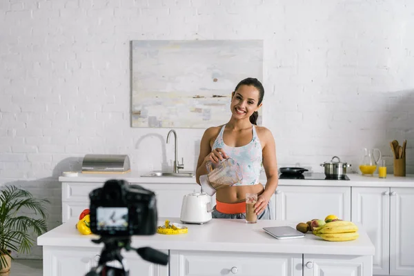 Selective focus of happy girl pouring smoothie in glass near digital camera — Stock Photo