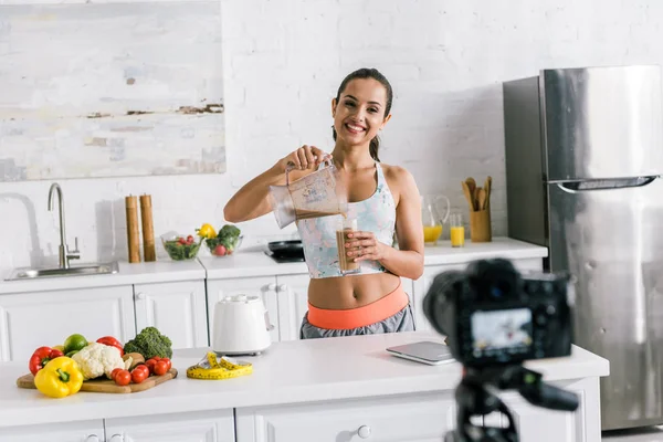 Selektiver Fokus einer glücklichen Frau, die Smoothie in die Nähe der Digitalkamera gießt — Stockfoto