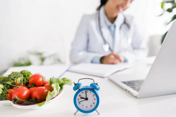 Cropped view of nutritionist near vegetables and alarm clock — Stock Photo