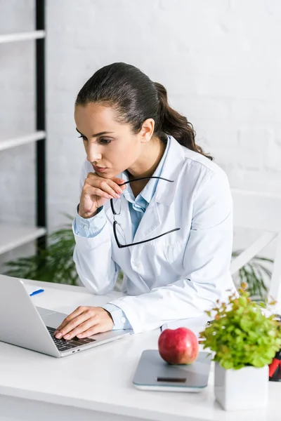 Nutritionniste attrayant en utilisant un ordinateur portable tout en tenant des lunettes — Photo de stock
