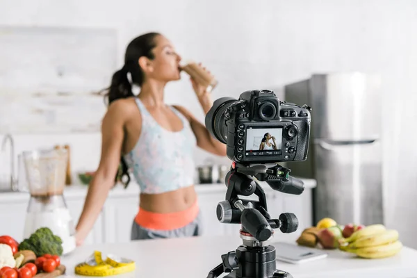 Foco seletivo da câmera digital com a menina bebendo smoothie perto de frutas e legumes na tela — Fotografia de Stock