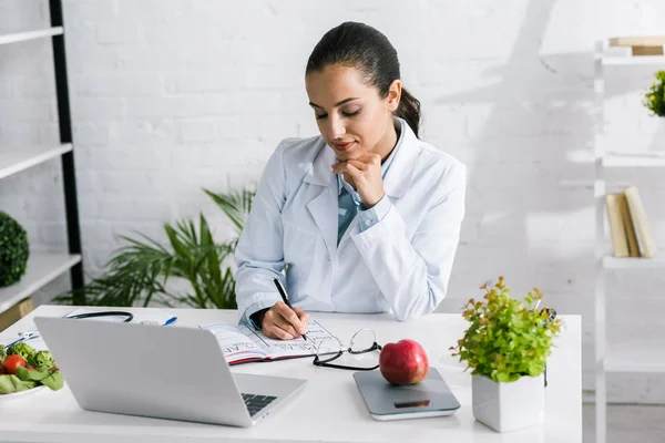 Attraktive Frau in weißem Mantel schreibt in Notizbuch neben Laptop und Gemüse — Stockfoto