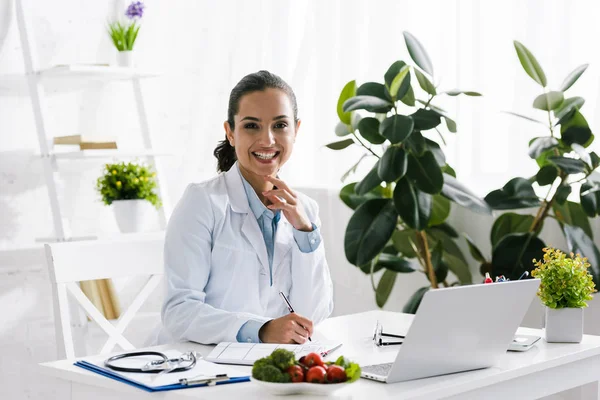 Glückliche Ernährungsberaterin im weißen Kittel neben Gemüse und Laptop — Stockfoto