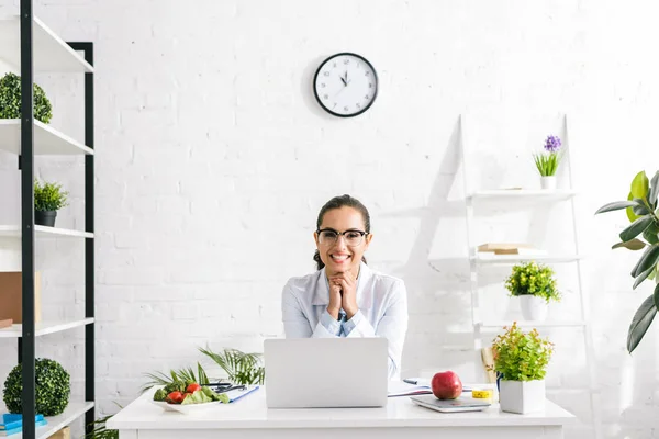 Cheerful nutritionist in white coat near vegetables and laptop — Stock Photo