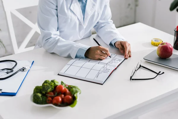 Cropped view of nutritionist writing in notebook near vegetables and apple — Stock Photo